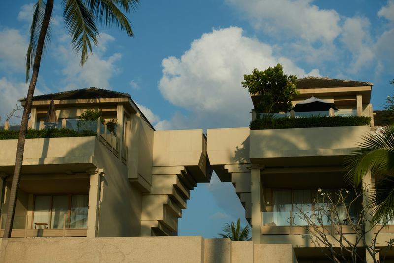 Deux petits immeubles résidentiels à toit-terrasse, séparés par un portail balinais fendu, avec des palmiers au premier plan